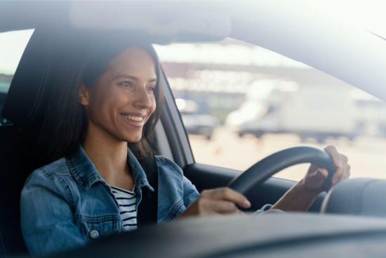 Mulher sorrindo ao dirigir um carro, com o volante nas mãos e expressão de tranquilidade, ao lado do banco do motorista, em ambiente externo com paisagem desfocada ao fundo.