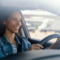 Mulher sorrindo ao dirigir um carro, com o volante nas mãos e expressão de tranquilidade, ao lado do banco do motorista, em ambiente externo com paisagem desfocada ao fundo.