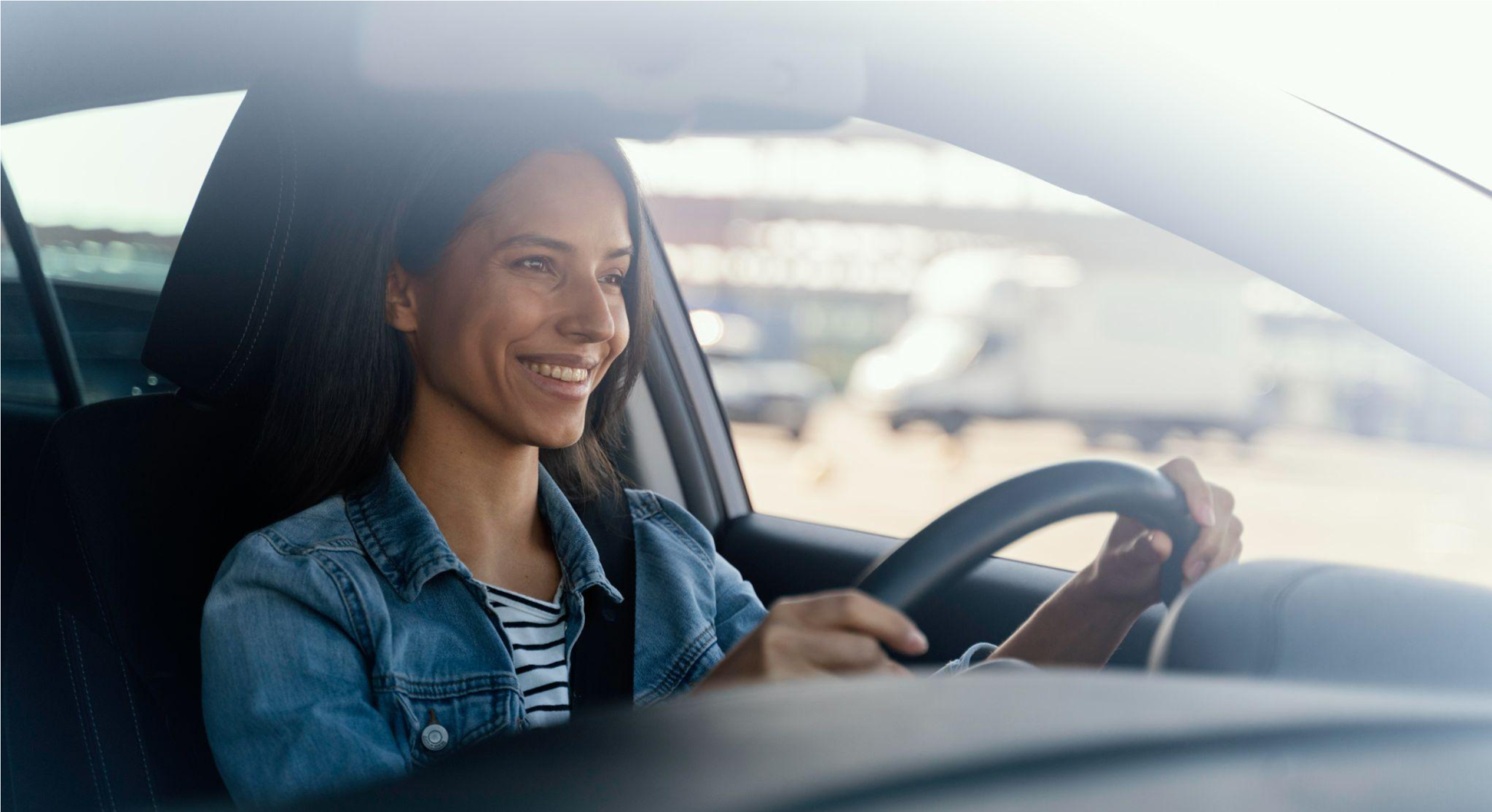 Mulher sorrindo ao dirigir um carro, com o volante nas mãos e expressão de tranquilidade, ao lado do banco do motorista, em ambiente externo com paisagem desfocada ao fundo.
