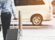 Homem com camisa azul e calça escura caminhando com mala de viagem grande e maleta de mão em um estacionamento, ao lado de um carro ao fundo, ambiente de transporte e viagem