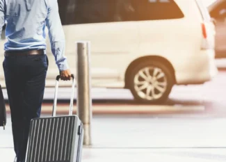 Homem com camisa azul e calça escura caminhando com mala de viagem grande e maleta de mão em um estacionamento, ao lado de um carro ao fundo, ambiente de transporte e viagem