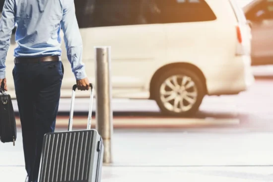 Homem com camisa azul e calça escura caminhando com mala de viagem grande e maleta de mão em um estacionamento, ao lado de um carro ao fundo, ambiente de transporte e viagem