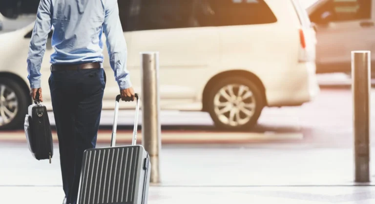 Homem com camisa azul e calça escura caminhando com mala de viagem grande e maleta de mão em um estacionamento, ao lado de um carro ao fundo, ambiente de transporte e viagem