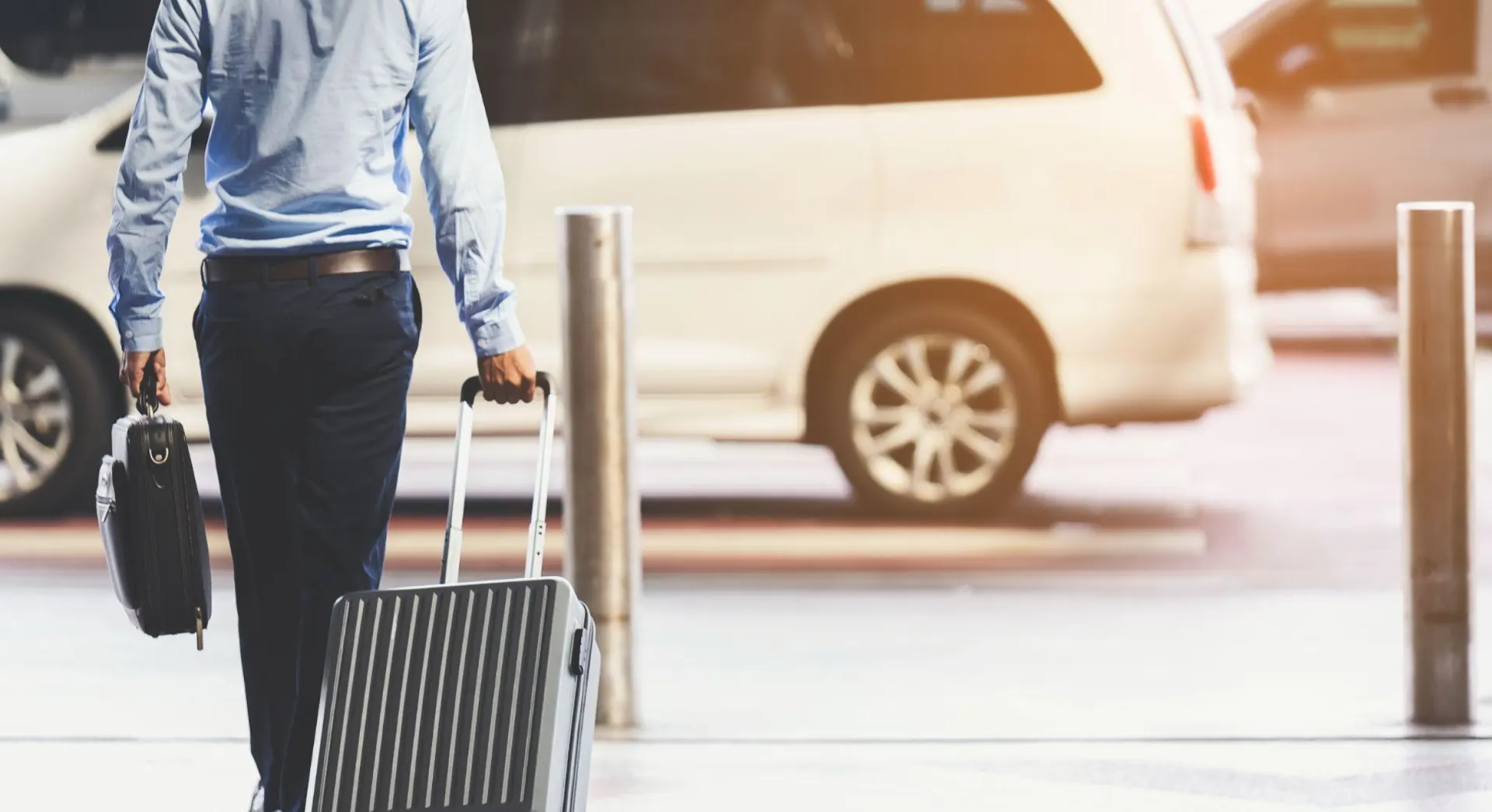 Homem com camisa azul e calça escura caminhando com mala de viagem grande e maleta de mão em um estacionamento, ao lado de um carro ao fundo, ambiente de transporte e viagem