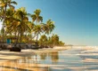 Praia tropical com fileira de coqueiros, areia clara e mar calmo ao amanhecer, com reflexos do céu na água e rochas na beira da costa ao fundo.