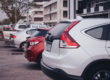 Fila de carros estacionados na rua, com veículos brancos e prata vistos de trás, em frente a um prédio ao fundo, com clima urbano ao entardecer.