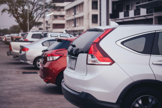 Fila de carros estacionados na rua, com veículos brancos e prata vistos de trás, em frente a um prédio ao fundo, com clima urbano ao entardecer.