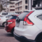 Fila de carros estacionados na rua, com veículos brancos e prata vistos de trás, em frente a um prédio ao fundo, com clima urbano ao entardecer.