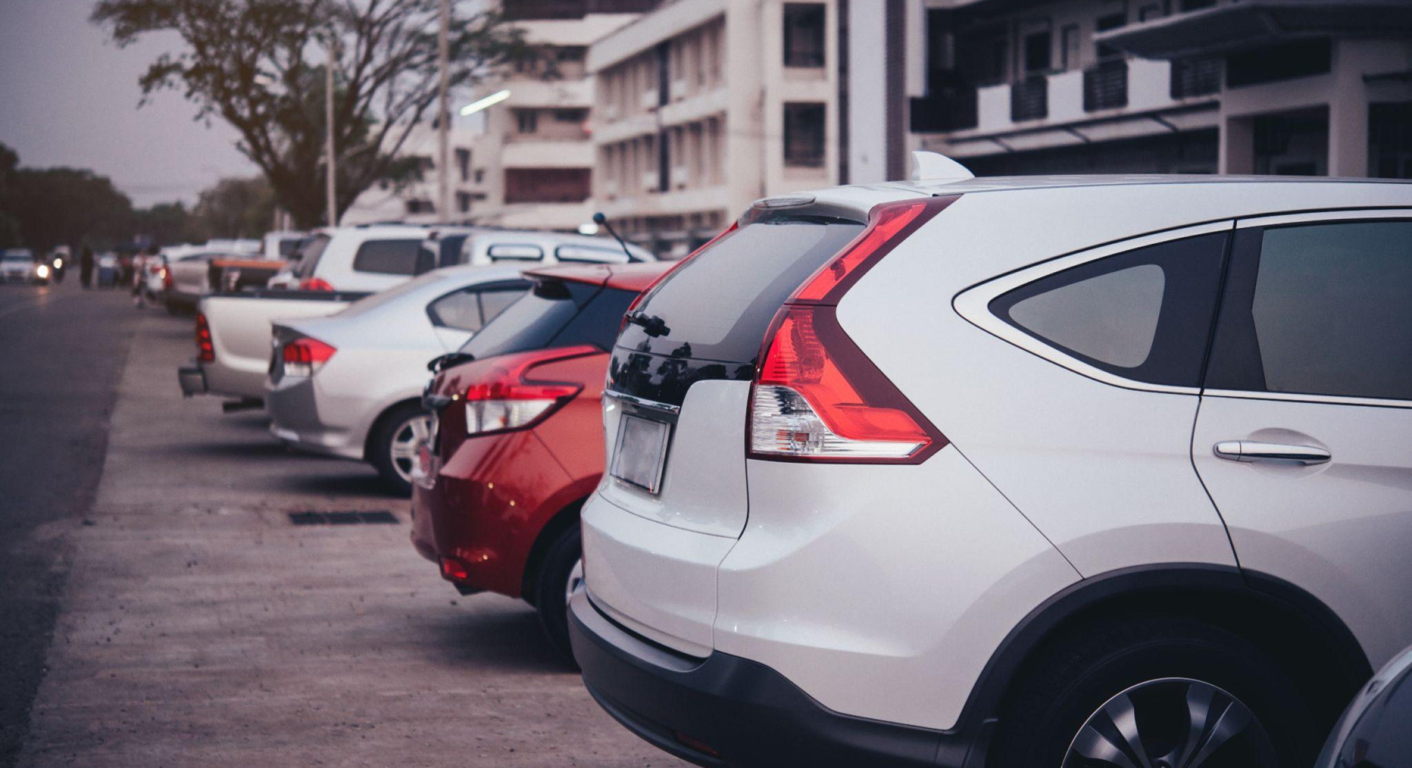 Fila de carros estacionados na rua, com veículos brancos e prata vistos de trás, em frente a um prédio ao fundo, com clima urbano ao entardecer.