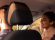 Casal dentro do carro durante um passeio, com o motorista ao volante e a passageira sorrindo ao lado pela janela, em clima de conversa e tranquilidade.