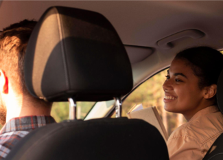 Casal dentro do carro durante um passeio, com o motorista ao volante e a passageira sorrindo ao lado pela janela, em clima de conversa e tranquilidade.