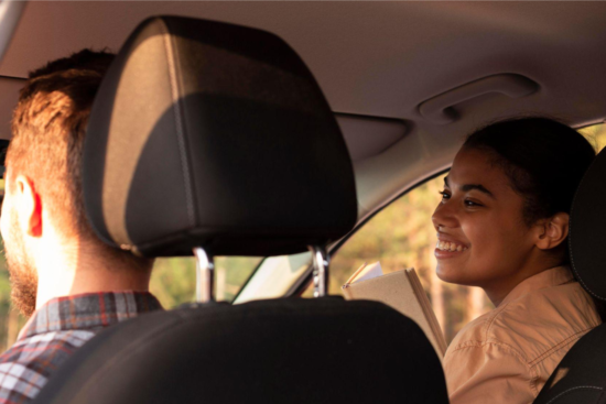 Casal dentro do carro durante um passeio, com o motorista ao volante e a passageira sorrindo ao lado pela janela, em clima de conversa e tranquilidade.