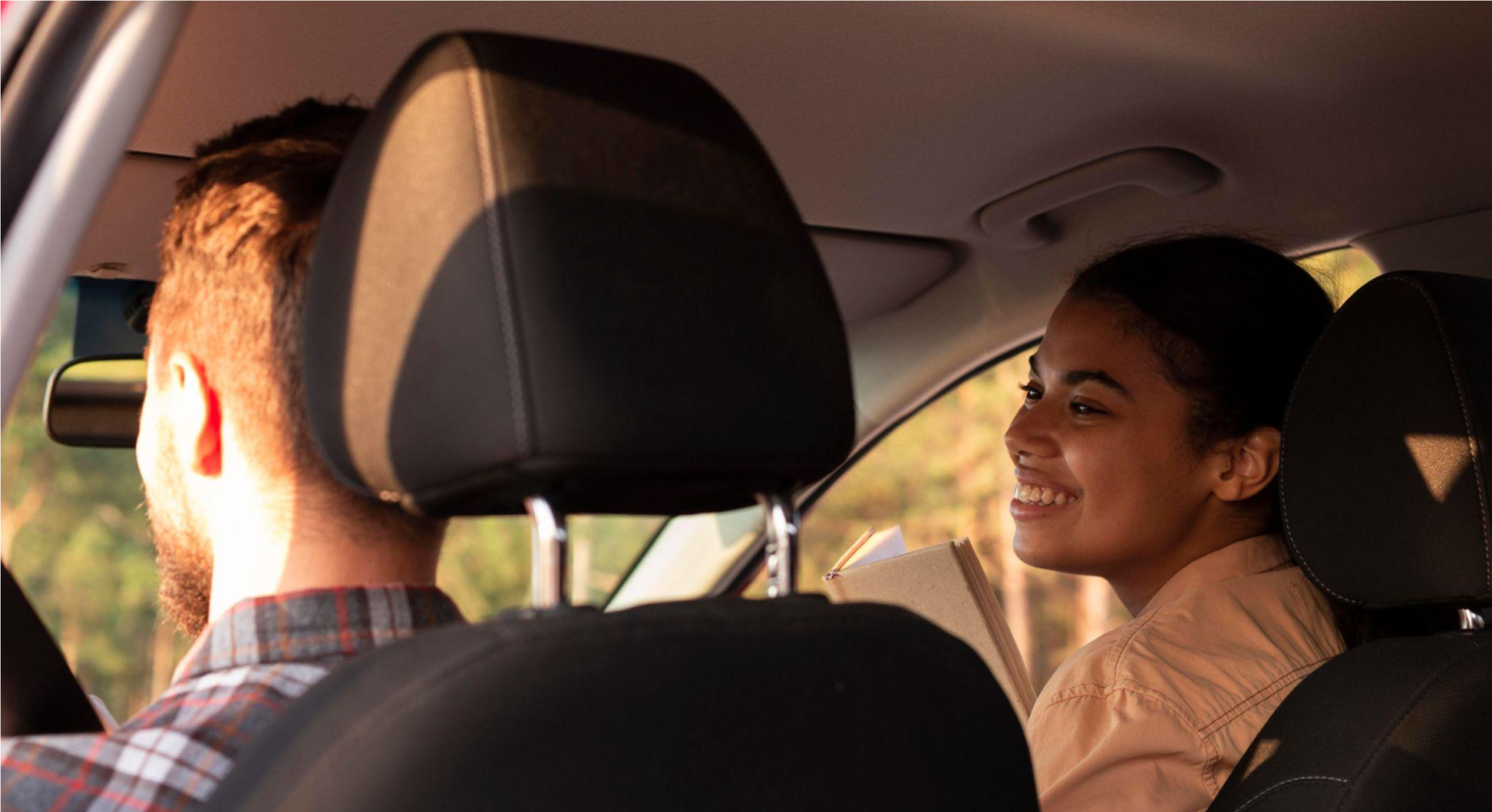 Casal dentro do carro durante um passeio, com o motorista ao volante e a passageira sorrindo ao lado pela janela, em clima de conversa e tranquilidade.