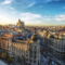 Vista aérea do centro de Madri, Espanha, com avenidas movimentadas e prédios históricos, incluindo a fachada de uma igreja e o horizonte com céu azul e nuvens.