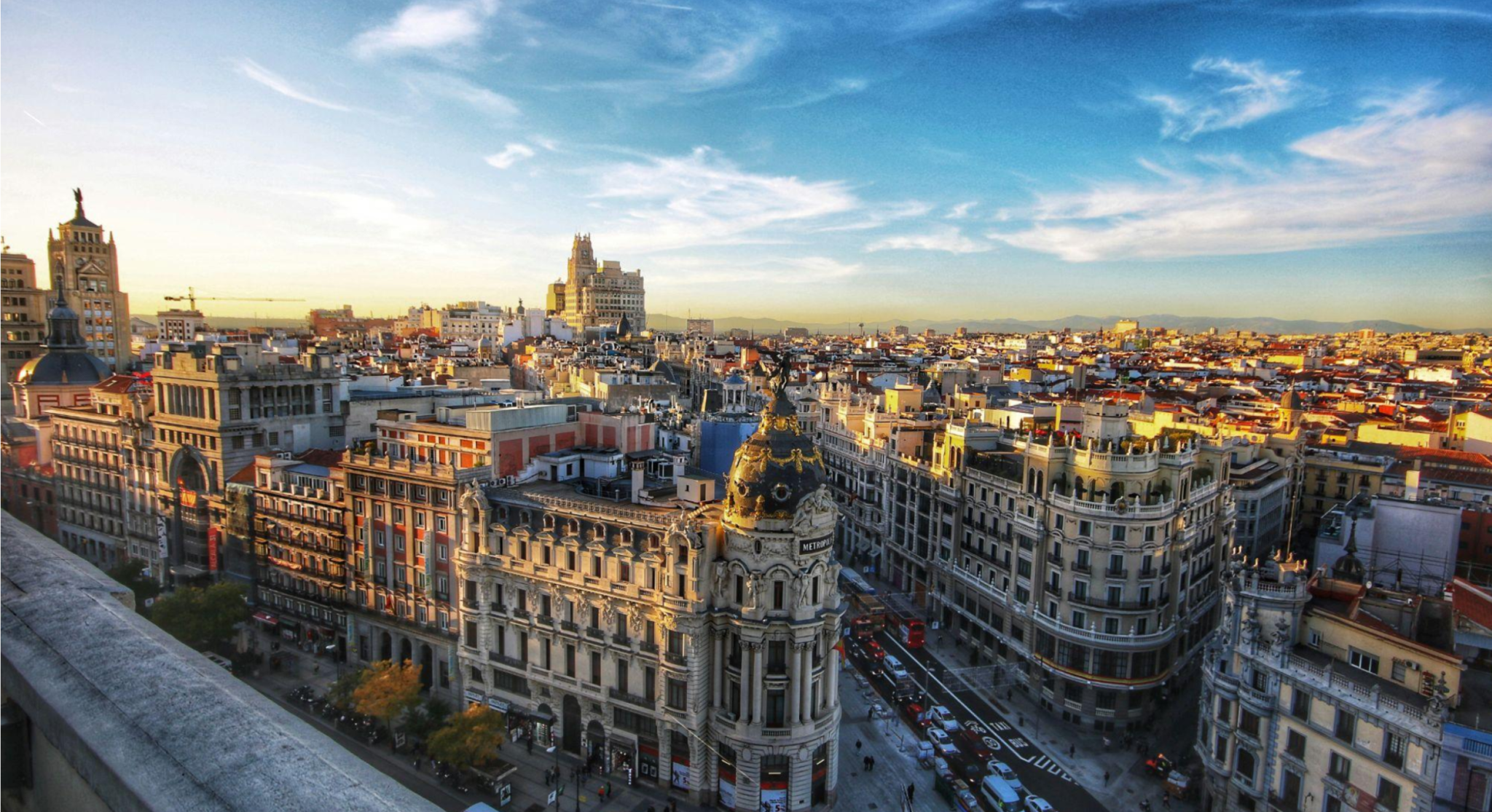Vista aérea do centro de Madri, Espanha, com avenidas movimentadas e prédios históricos, incluindo a fachada de uma igreja e o horizonte com céu azul e nuvens.