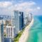 Vista aérea da orla da praia de Miami com arranha-céus e edifícios residenciais na cidade, mar azul e faixa de areia ao lado de torres altas sob céu parcialmente nublado.