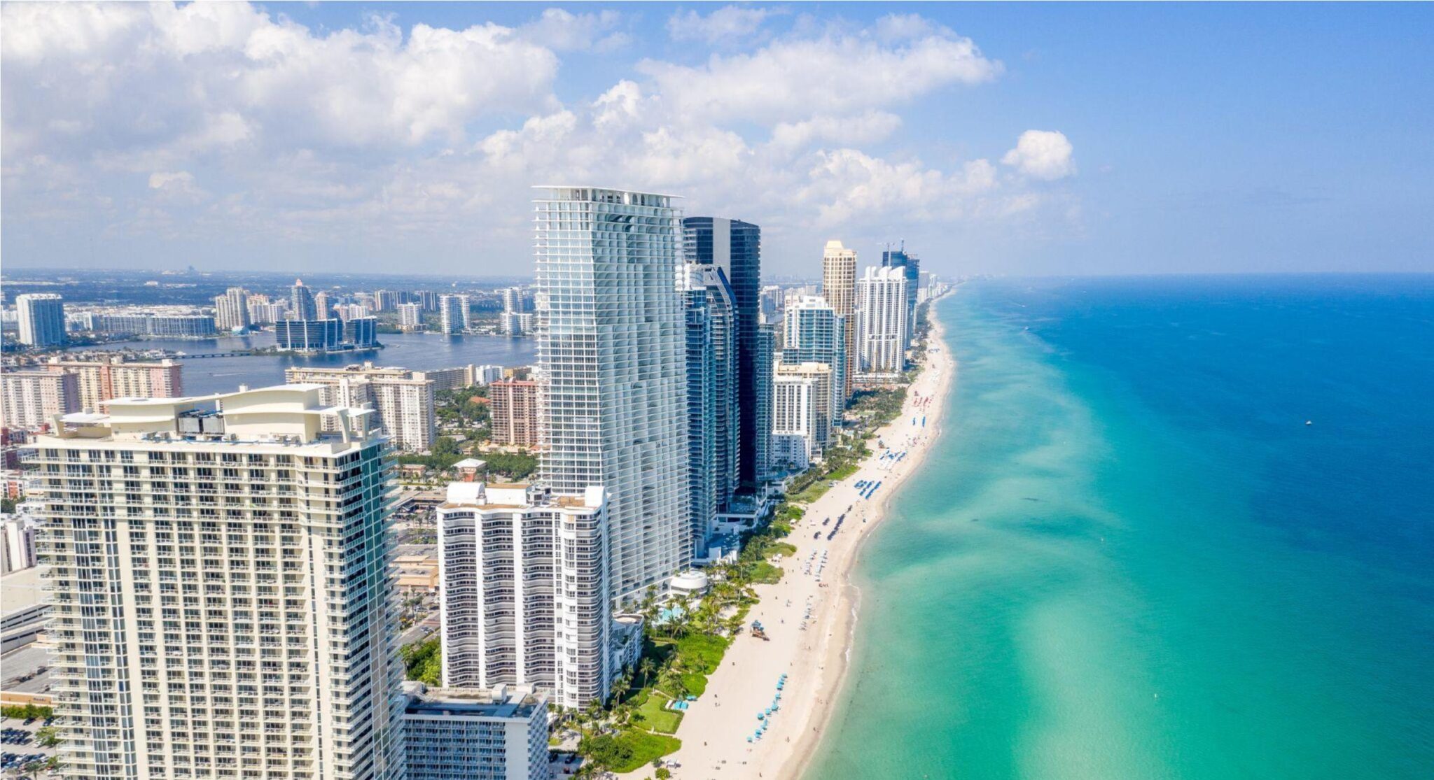 Vista aérea da orla da praia de Miami com arranha-céus e edifícios residenciais na cidade, mar azul e faixa de areia ao lado de torres altas sob céu parcialmente nublado.