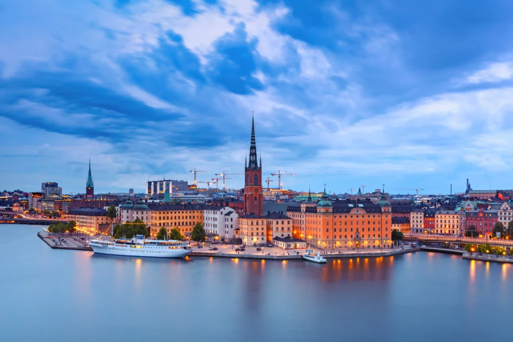 Vista panorâmica do centro histórico de Estocolmo ao entardecer, com o Rathaus (Câmara Municipal) ao fundo, prédios coloridos e o porto com barcos e navios refletidos na água calma sob céu azul com nuvens
