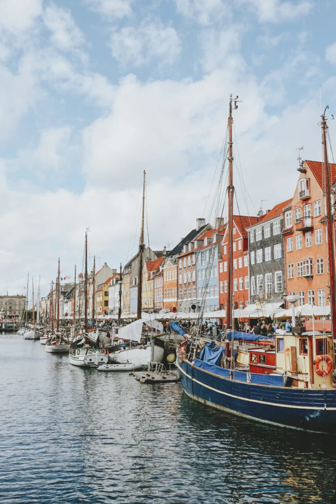 Vista do porto com barcos atracados e casas coloridas com telhados de cerâmica em tom vermelho, amarelo e branco, céu azul com nuvens e mastro de embarcações