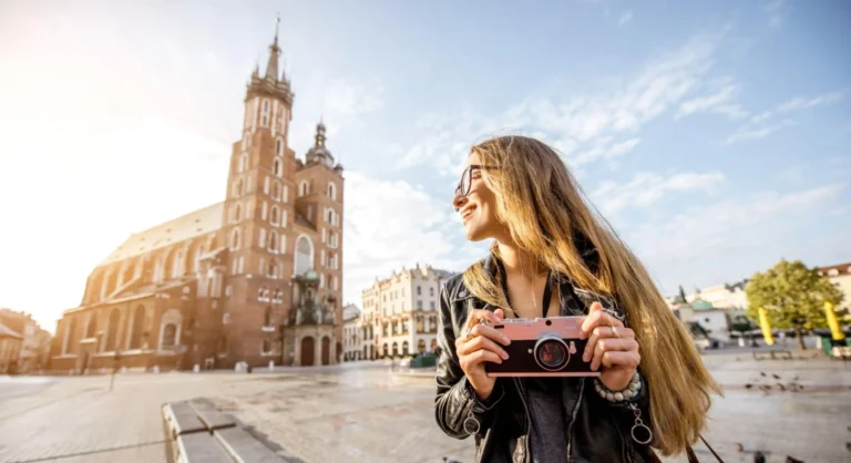 Mulher ao ar livre em um passeio turístico segura uma câmera fotográfica e fotografa em frente à catedral de Cracóvia, com céu azul e luz do pôr do sol