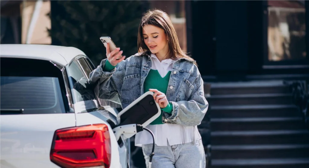A woman stands beside a eletric car in a parking lot, holding a charging cable and checking her phone while connecting it to charge the electric vehicle.