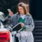 A woman stands beside a eletric car in a parking lot, holding a charging cable and checking her phone while connecting it to charge the electric vehicle.