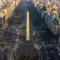 Vista aérea de um obelisco em avenida urbana com prédios altos ao redor, carros e faixas de trânsito, em uma cidade movimentada.