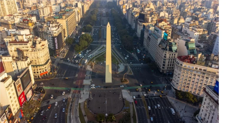 Vista aérea de um obelisco em avenida urbana com prédios altos ao redor, carros e faixas de trânsito, em uma cidade movimentada.