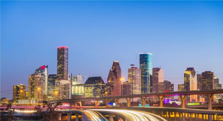 Vista noturna em Houston Texas, com arranha-céus iluminados e um viaduto sobre um rio, com rastros de luz de carros em movimento ao entardecer