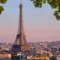 Torre Eiffel ao pôr do sol em Paris, vista panorâmica da cidade com edifícios ao fundo e folhas verdes enquadrando a paisagem.