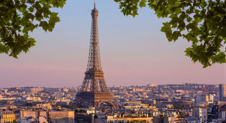 Torre Eiffel ao pôr do sol em Paris, vista panorâmica da cidade com edifícios ao fundo e folhas verdes enquadrando a paisagem.