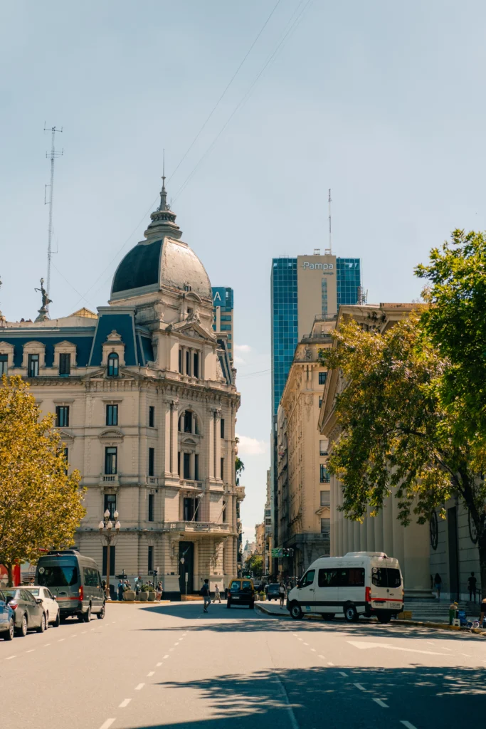 Avenida em Buenos Aires com prédios históricos e modernos ao fundo, com cúpula monumental de igreja e ônibus circulando na via em dia ensolarado