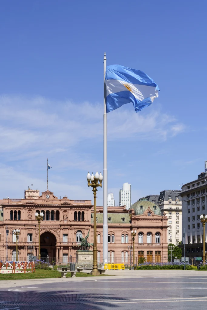Bandeira argentina ao vento em frente a prédios históricos na cidade, com céu azul e nuvens leves ao fundo.