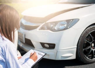 Persona junto a un auto blanco, revisando los documentos del seguro LDW y completando un formulario al aire libre, con detalles del neumático y el faro visibles.