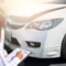 Persona junto a un auto blanco, revisando los documentos del seguro LDW y completando un formulario al aire libre, con detalles del neumático y el faro visibles.
