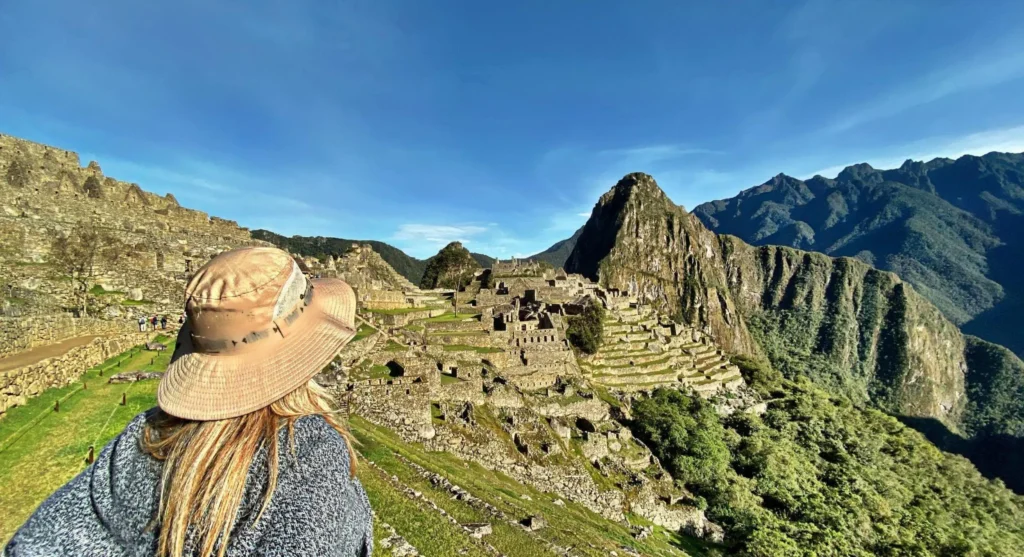 Mulher de chapéu de palha olhando para Machu Picchu nas montanhas do Peru, com ruínas antigas em meio à vegetação e céu azul nítido.