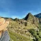Mulher de chapéu de palha olhando para Machu Picchu nas montanhas do Peru, com ruínas antigas em meio à vegetação e céu azul nítido.