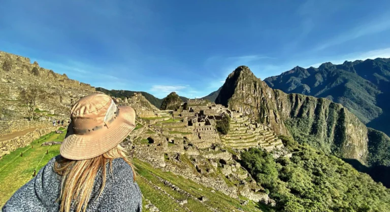 Mulher de chapéu de palha olhando para Machu Picchu nas montanhas do Peru, com ruínas antigas em meio à vegetação e céu azul nítido.
