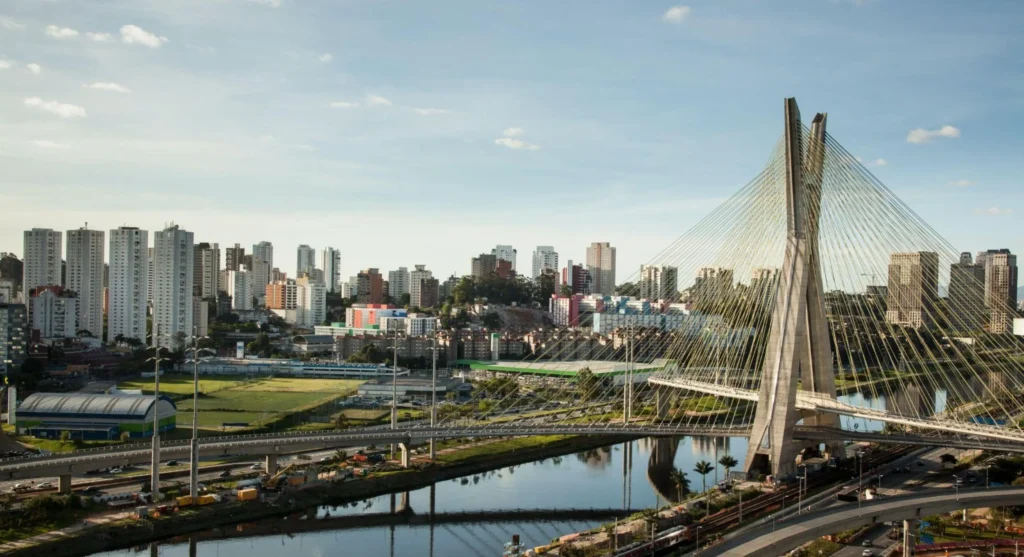 Vista panorâmica da Ponte Estaiada Octávio Frias de Oliveira em São Paulo sobre o rio, com arranha-céus ao fundo e tráfego em avenidas na cidade ao amanhecer.