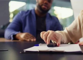 Pessoas em reunião, com uma das pessoas assinando documentos sobre uma mesa, enquanto outras discutem ao fundo, em ambiente corporativo.