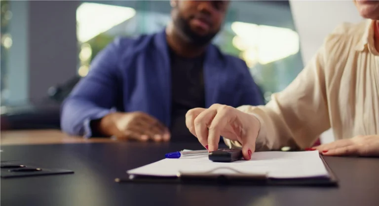 Pessoas em reunião, com uma das pessoas assinando documentos sobre uma mesa, enquanto outras discutem ao fundo, em ambiente corporativo.
