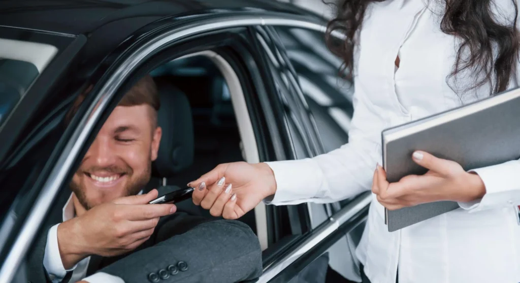 Atendimento em carro: motorista sorridente recebe as mãos de um consultor que entrega a chave do carro, sugerindo suporte ou verificação de informações durante a entrega.