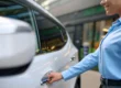 Mulher de blusa azul no pátio de uma oficina ou estacionamento, segurando a chave e abrindo a porta de um carro branco, mostrando a ação de atendimento e acesso ao veículo.
