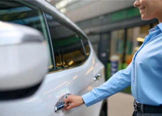Mulher de blusa azul no pátio de uma oficina ou estacionamento, segurando a chave e abrindo a porta de um carro branco, mostrando a ação de atendimento e acesso ao veículo.