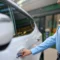 Mulher de blusa azul no pátio de uma oficina ou estacionamento, segurando a chave e abrindo a porta de um carro branco, mostrando a ação de atendimento e acesso ao veículo.