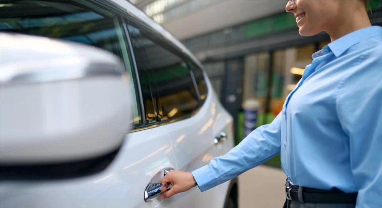Mulher de blusa azul no pátio de uma oficina ou estacionamento, segurando a chave e abrindo a porta de um carro branco, mostrando a ação de atendimento e acesso ao veículo.